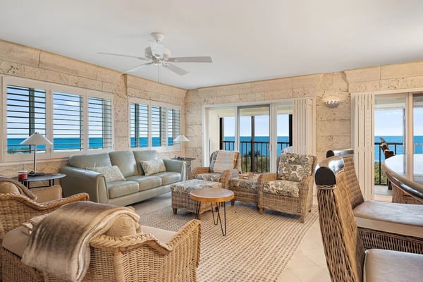 Beachfront living room with wicker furniture, stone walls, ocean views through shuttered windows and glass doors.